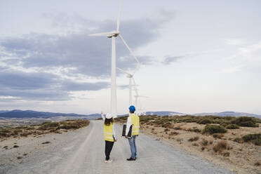 Engineers standing on dirt road looking at wind turbines at wind farm - EBBF06363