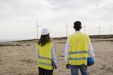 Colleagues wearing reflective clothing looking at wind turbines standing at wind farm - EBBF06369
