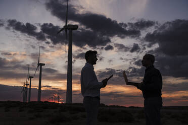 Engineers discussing with each other holding laptop at wind farm on sunset - EBBF06376