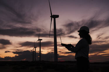 Silhouette of engineer working on laptop by wind turbines at wind farm - EBBF06390