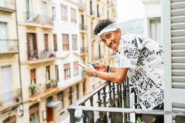 Seitenansicht eines fröhlichen ethnischen Mannes, der auf dem Balkon steht und auf seinem Smartphone surft, während er seinen Morgenkaffee genießt und in die Kamera schaut - ADSF37659