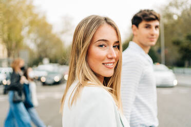 Side view of content stylish couple holding hands and crossing road while walking in city and looking at camera - ADSF37664