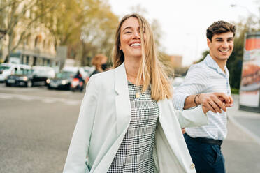 Side view of content stylish couple holding hands and crossing road while walking in city and looking away - ADSF37665