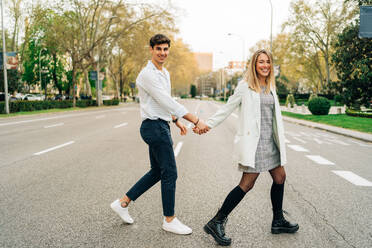 Side view of content stylish couple holding hands and crossing road while walking in city and looking at camera - ADSF37670