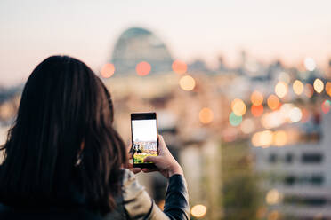 Rückansicht einer nicht erkennbaren Frau, die den Bildschirm ihres Handys berührt, während sie ein Foto von der Stadt am Abend macht - ADSF37699