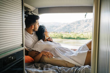 Zartes spanisches Paar auf Reisen, das sich auf dem Bett im Wohnwagen entspannt, während es die Reise genießt und nach draußen schaut - ADSF37707