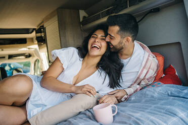 Zartes hispanisches Paar auf Reisen lacht zusammen auf dem Bett und kuschelt im Wohnwagen, während es die Reise genießt - ADSF37710