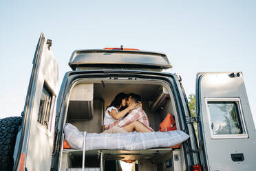 Low angle side view of traveling couple sitting on bed in camper and embracing and kissing during summer trip - ADSF37712