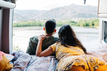 Back view of unrecognizable romantic couple enjoying landscape of green hills and river while resting together in camper van during summer trip in nature - ADSF37723
