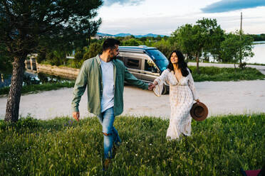 Glückliches junges Paar, das sich an den Händen hält, während es gemeinsam auf grünem Gras in der Nähe eines geparkten Wohnmobils während einer Sommerreise in der Natur spazieren geht - ADSF37727