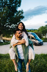 Young man giving piggyback ride to happy girlfriend while resting together on green grassy meadow in hilly countryside in summer day - ADSF37728