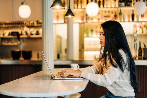 Junge ethnische Frau, die am Tisch in der Cafeteria auf einem Netbook surft - ADSF37731