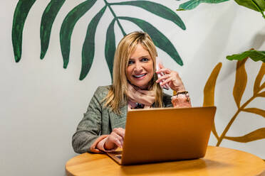 Erwachsene fröhliche weibliche Führungskraft in formeller Kleidung, die mit einem Handy vor einem Netbook spricht, während sie in die Kamera in der Nähe einer ornamentalen Wand schaut - ADSF37746