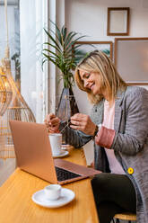 Erwachsener lächelnder Unternehmer mit Tasse Heißgetränk und Netbook am Tisch im Café sitzend - ADSF37748