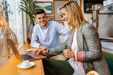 Seitenansicht einer lächelnden Business-Trainerin, die sich mit einem Mitarbeiter unterhält, während sie mit einem Netbook am Tisch in der Cafeteria sitzen und sich gegenseitig ansehen - ADSF37751