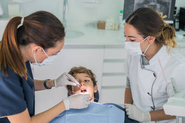Von oben weiblicher Arzt und Krankenschwester mit zahnärztlichen Werkzeugen Überprüfung und Behandlung der Zähne des Jungen während der Arbeit in der zeitgenössischen Klinik - ADSF37772