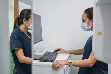 Side view of woman in medical uniform looking at teeth X ray on computer monitor while working in lab of contemporary dental clinic - ADSF37779