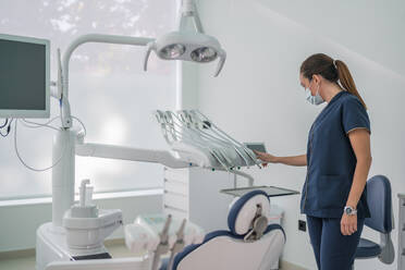 Side view of woman in medical uniform and mask adjusting tools settings while standing in light dental office before curing procedure - ADSF37783