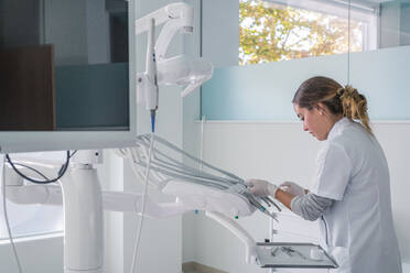 Seitenansicht einer Frau in medizinischer Uniform, die vor der Behandlung in einer hellen Zahnarztpraxis steht und die Einstellungen für die Instrumente vornimmt - ADSF37784