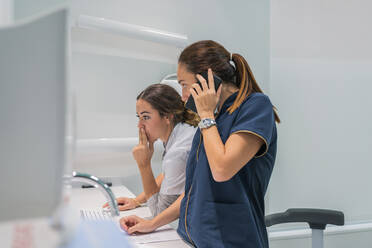 Seitenansicht eines Mediziners, der im Labor eines modernen Krankenhauses Daten auf einem modernen Computer durchsucht und dabei mit einem Mobiltelefon spricht - ADSF37787