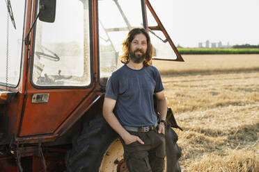 Lächelnder Landwirt mit Händen in den Taschen, der sich auf einem Bauernhof an einen Traktor lehnt - EKGF00005