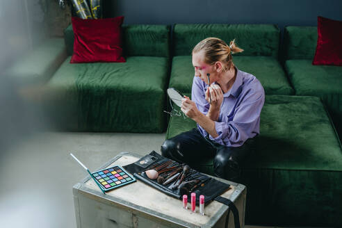 Man applying make-up looking in hand mirror sitting on sofa - YTF00015