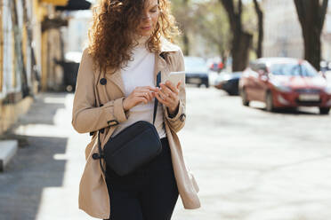 Woman with shoulder bag using mobile phone at sidewalk on sunny day - OYF00727