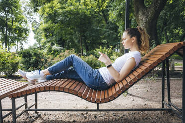 Frau, die ein Mobiltelefon benutzt und sich auf einem Liegestuhl im Park entspannt - OYF00732