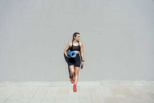Junge Frau mit Trainingsmatte an grauer Wand lehnend - OIPF02403