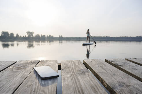Mobile phone on jetty with woman doing standup paddleboarding in background at sunset - EYAF02159