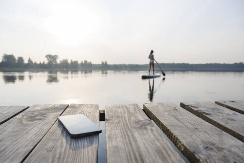 Mobiltelefon auf einem Steg mit einer Frau, die im Hintergrund bei Sonnenuntergang Standup-Paddleboarding betreibt - EYAF02159