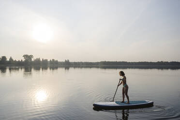 Frau beim Standup-Paddleboarding im See bei Sonnenuntergang - EYAF02160