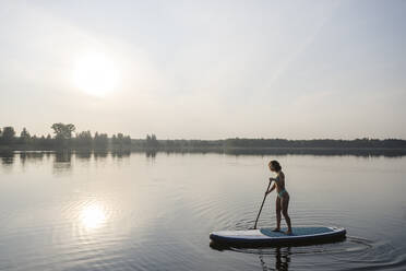 Woman doing standup paddleboarding in lake at sunset - EYAF02160