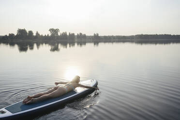 Woman paddling with hand lying on paddleboard at sunset - EYAF02162