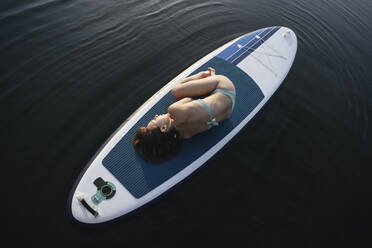 Woman lying in embryo pose on paddleboard over water - EYAF02163