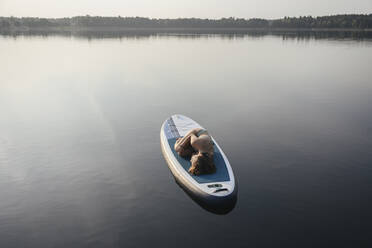 Woman lying in embryo pose on paddleboard at lake - EYAF02165