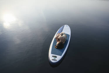 Woman lying in embryo pose on paddleboard at sunset - EYAF02166