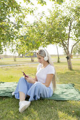 Young woman with wireless headphones using smart phone sitting in park - LESF00166