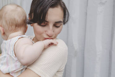 Mother kissing baby girl's little hand in front of wall - IHF01224