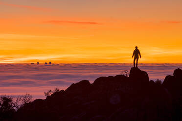 Rückansicht der Silhouette eines männlichen Touristen, der auf einem Felsen steht und den schönen Sonnenuntergang über einer mit Nebel und Wolken bedeckten Stadt während einer Reise in Spanien genießt - ADSF37808
