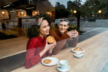 Through glass of cheerful young multiracial couple in love sitting at wooden counter and enjoying delicious pastry and coffee in cozy cafeteria - ADSF37831
