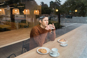 Young male sitting at wooden counter near window and looking away pensively while enjoying aromatic coffee and croissant in modern cafeteria - ADSF37832