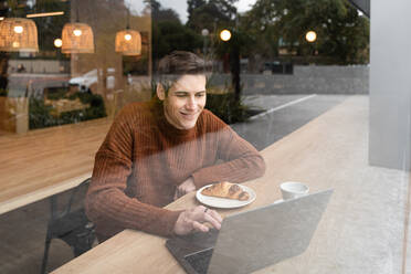 Durch das Fenster eines glücklichen jungen männlichen Studenten, der mit einem Laptop und einer Tasse Kaffee mit Croissant am Tisch sitzt, während er seine Zeit in einer modernen Cafeteria verbringt - ADSF37833