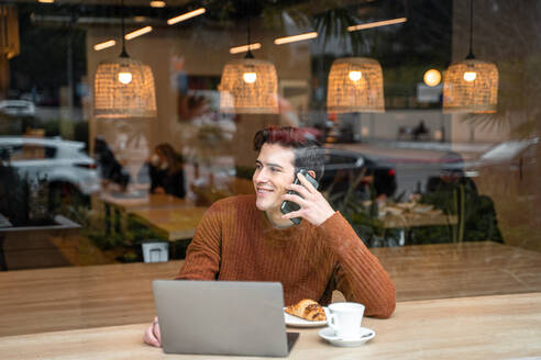 Durch das Fenster eines glücklichen jungen männlichen Studenten, der an einem Tisch mit Laptop und einer Tasse Kaffee mit Croissant sitzt und mit seinem Handy telefoniert, während er seine Zeit in einer modernen Cafeteria verbringt - ADSF37834
