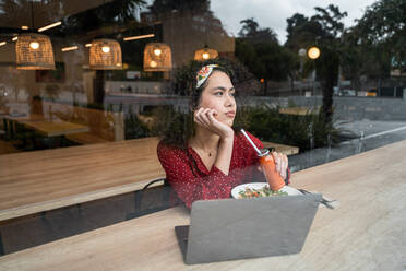 Durch das Glas einer jungen Bloggerin, die mit einer gesunden Mahlzeit am Tisch sitzt und einen Laptop benutzt, während sie Zeit in einem modernen Restaurant verbringt - ADSF37838