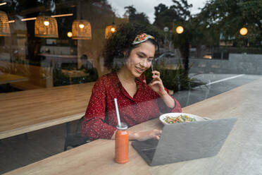 Through glass of positive young female blogger sitting at table with healthy meal and using laptop and talking phone while spending time in modern restaurant - ADSF37839
