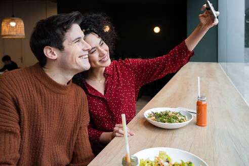 Fröhliches junges gemischtrassiges Paar sitzt am Tresen mit gesundem vegetarischem Salat und Getränken und macht ein Selfie mit dem Smartphone beim gemeinsamen Frühstück in einem modernen Restaurant - ADSF37840