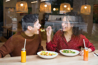 Durch Glas lachende ethnische Frau füttert Freund mit köstlichem vegetarischem Salat beim gemeinsamen gesunden Essen am Tresen eines modernen Restaurants - ADSF37841