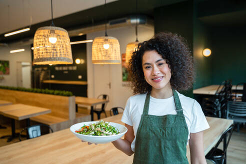 Delighted ethnic waitress in apron standing in empty cafe with plate of tasty healthy salad and looking at camera - ADSF37846