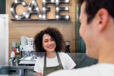 Hospitable ethnic female barista in apron standing at counter in cafe and talking to male client - ADSF37847